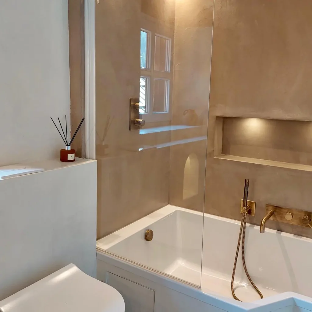 Salle de bain moderne avec baignoire et murs en béton ciré beige, équipée de robinetterie en laiton doré et niches éclairées pour un style élégant et minimaliste.