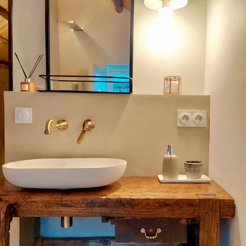 Lavabo moderne en céramique blanche sur un meuble en bois rustique avec un mur en béton ciré et des robinets en laiton doré, combinant modernité et charme ancien. 