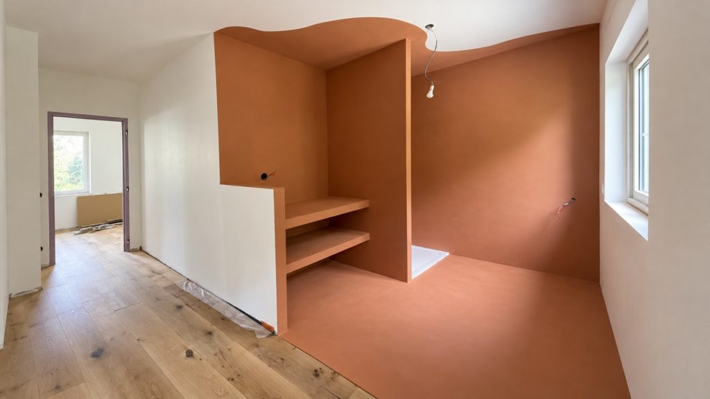 Salle d’eau aux murs, étagères maçonnées et sol en béton ciré terracotta, plafond découpé en vague, à côté d’un couloir en parquet de chêne — chantier BTONBYSANDRA, béton ciré mural en PACA