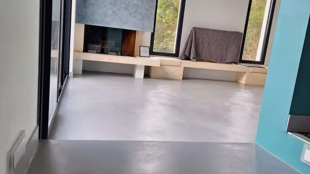 Séjour de rez-de-chaussée au sol en béton ciré gris continu sans joint, cheminée et banquette en pierre, grandes baies vitrées — réalisation BTONBYSANDRA, artisane Marius Aurenti en PACA